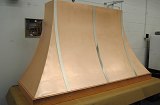 Wall-mounted copper alloy kitchen hood with stainless steel trim and rivets. Complete fabrication – cut, formed, rolled, corner welded with blending and DA finished prior to final antique finish applied.