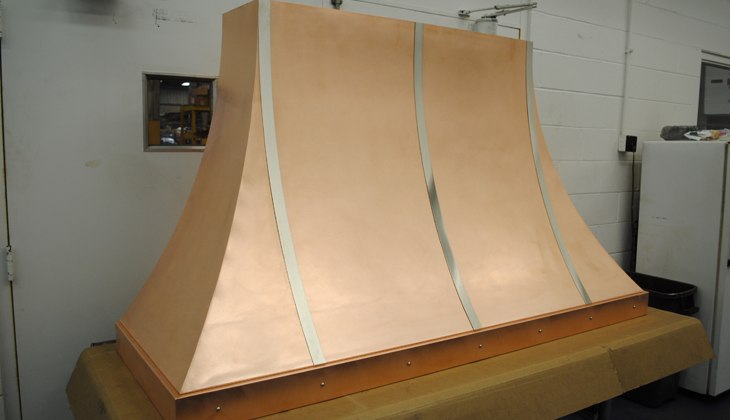 Wall-mounted copper alloy kitchen hood with stainless steel trim and rivets. Complete fabrication – cut, formed, rolled, corner welded with blending and DA finished prior to final antique finish applied.